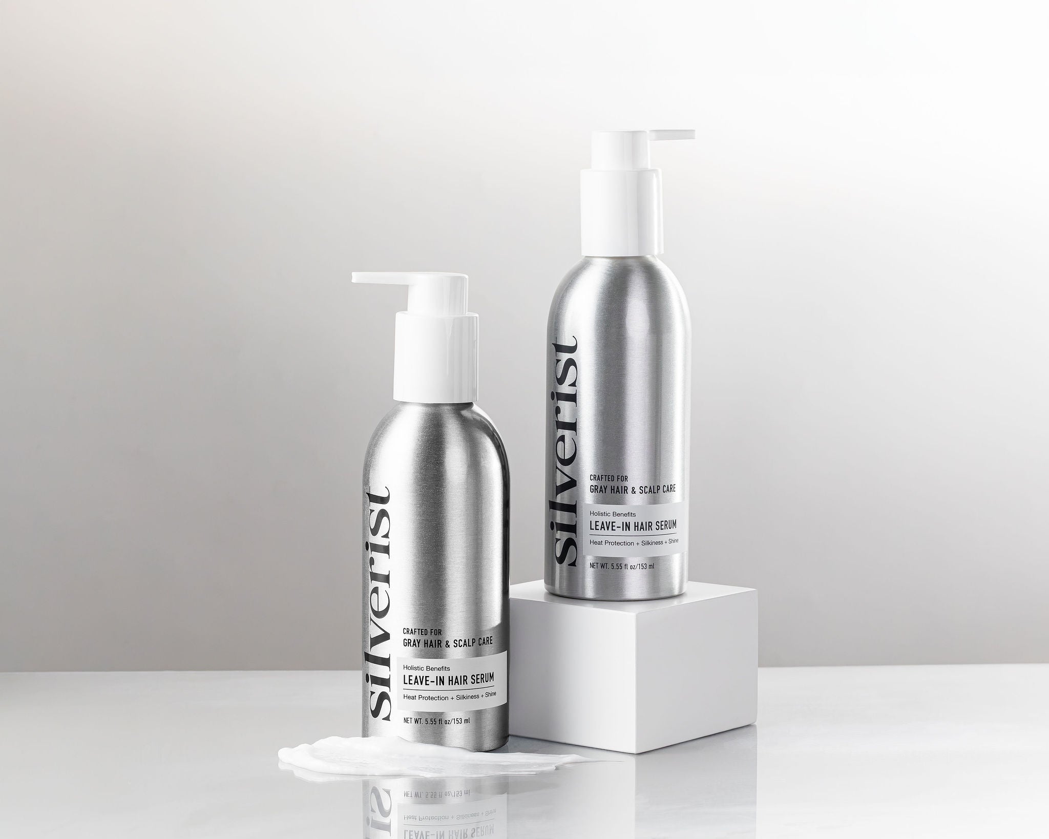 Two silver pump bottles of Silverist Holistic Benefits Leave-In Serum with a white pedestal against a light gray background.